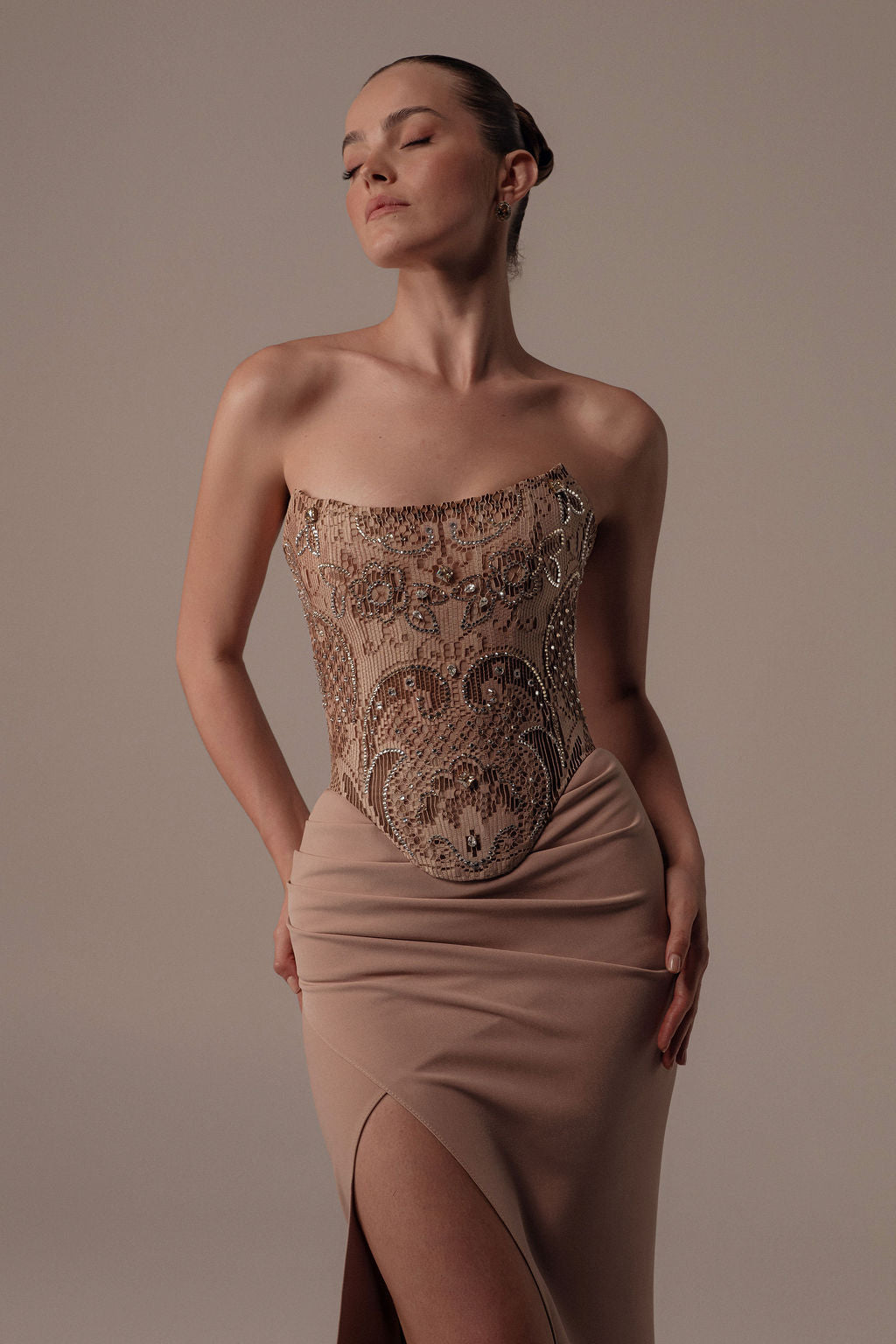 אישה פוזלת בחינניות בשמלת Dana מקולקציית הערב 2026 של דרור קונטנטו, עם מחשוף סטרפלס משובץ חרוזים בצבע גוף וחצאית בצבע בז' עם חתך גבוה. היא עומדת עם עיניים עצומות וידיים על המותניים על רקע ניטרלי.