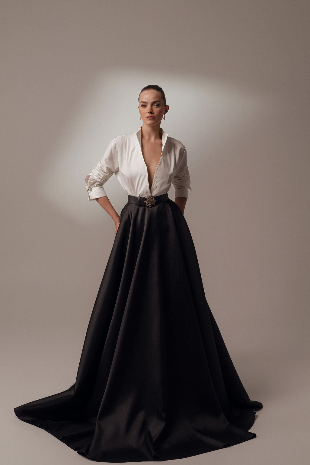 אישה עומדת על רקע אחיד לבושה בשמלת Elyn מקולקציית ערב 2026 של דרור קונטנטו — חולצה לבנה מפוארת עם שרוולים ארוכים, מחשוף עמוק וחצאית שחורה ארוכה עם מותן גבוהה. שיערה אסוף לאחור.