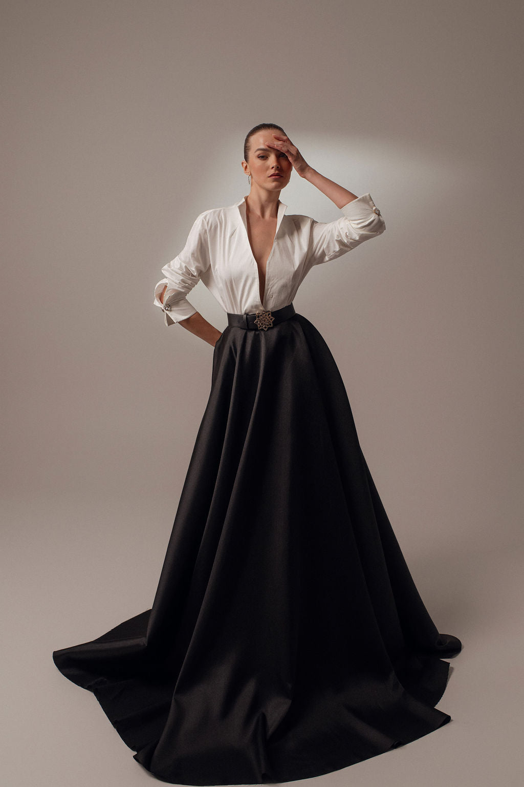 אישה מצטלמת על רקע אחיד כשהיא לובשת את שמלת Elyn מקולקציית Evening 2026 — חצאית שחורה ארוכה ומלאה עם מותניים גבוהים וחולצה לבנה עם מחשוף V עמוק. היא עומדת עם יד אחת על המותן והשנייה מורמת אל המצח.
