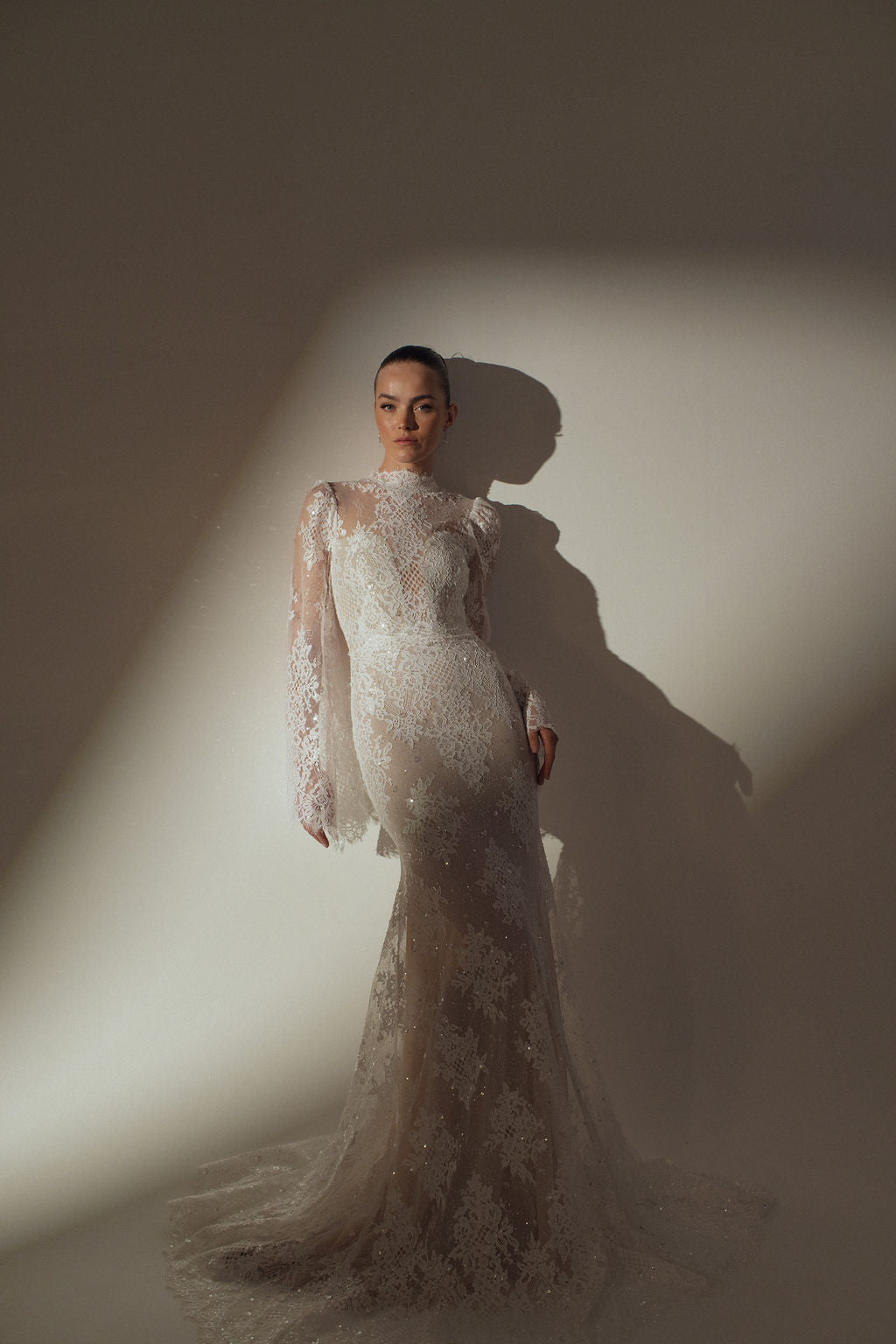 אישה לבושה בשמלת קירה שקופה עם שרוולים ארוכים מקולקציית ערב 2026 עומדת מול קיר חלק, מוארת באור רך עם צללים דרמטיים מאחוריה.