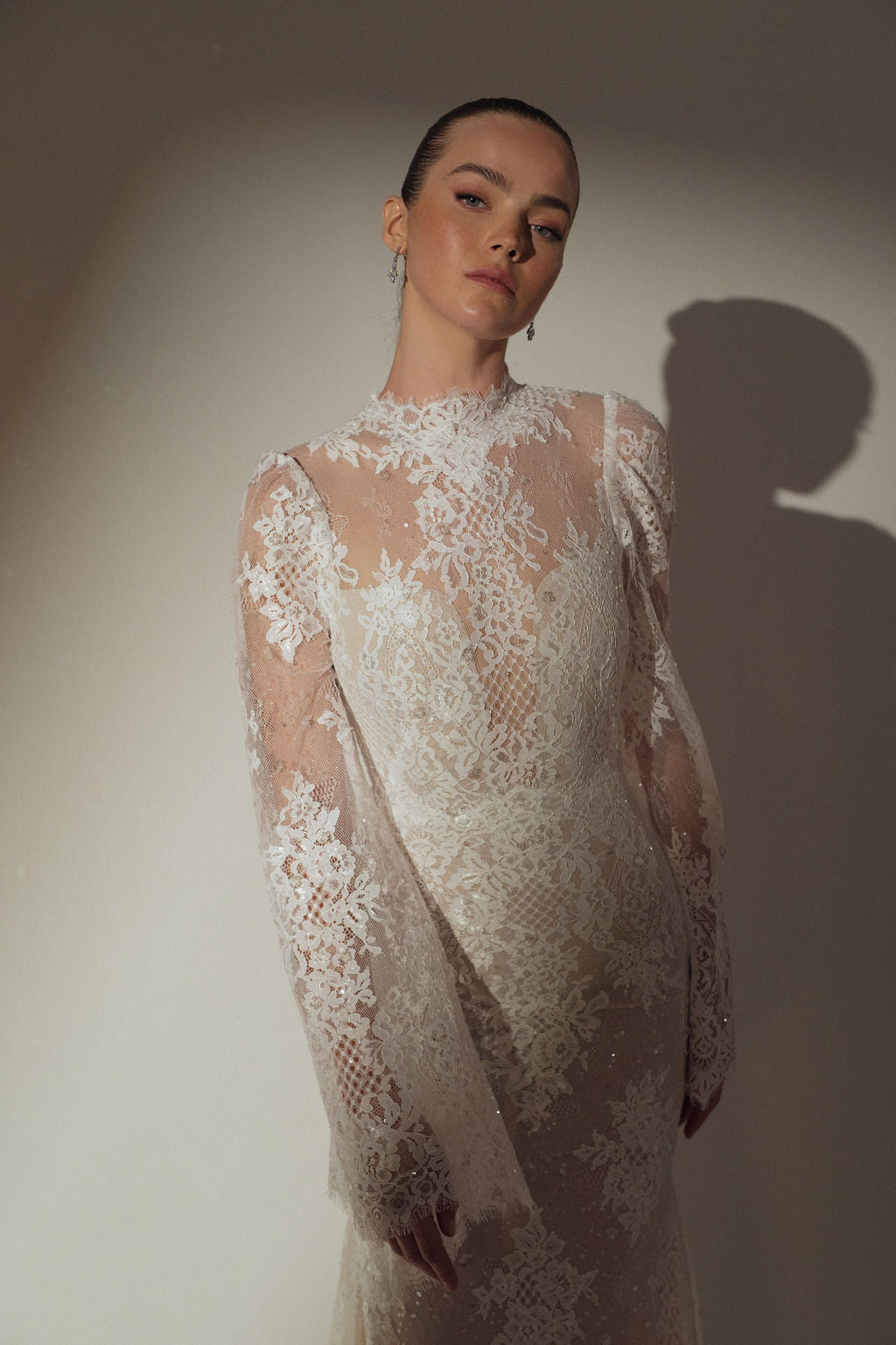 אישה בשמלת Kira מקולקציית ערב 2026 של דרור קונטנטו עומדת על רקע ניטרלי, מביטה בביטחון למצלמה, שיערה אסוף לאחור ותאורה רכה מטילה צללים מאחוריה.