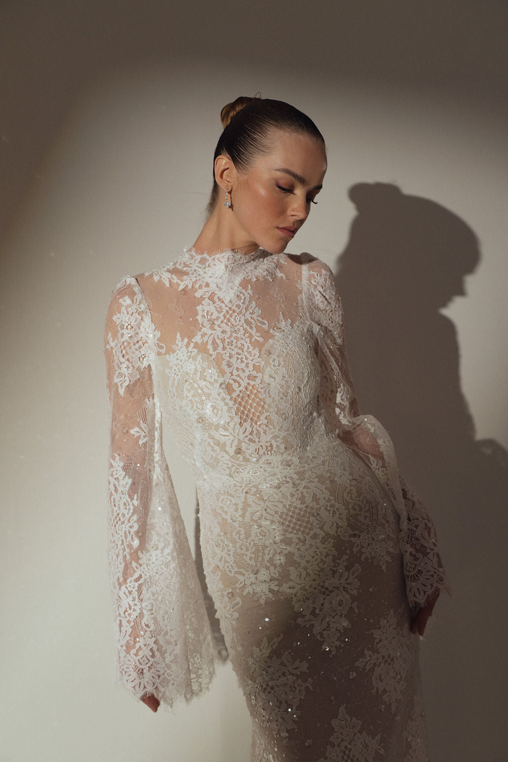 אישה לבושה בשמלת Kira מקולקציית ערב 2026 של דרור קונטנטו עומדת מול קיר חלק. השמלה השקופה עם שרוולים ארוכים מעוטרת בדוגמאות מורכבות, והיא מביטה למטה בשלווה, שיערה אסוף בקוקו חלק.