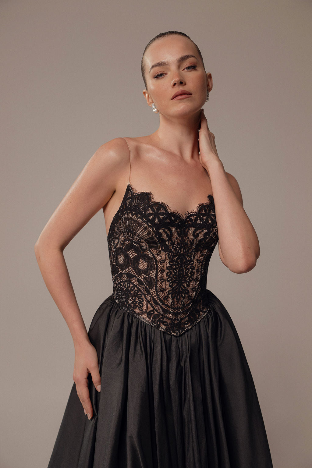 אישה עם שיער אסוף לאחור מדגמנת את שמלת Luna מקולקציית Evening 2026, עם מחוך תחרה וחצאית סאטן מלאה, ומצטלמת על רקע בז' עם יד אחת על צווארה והשנייה על מותנה.