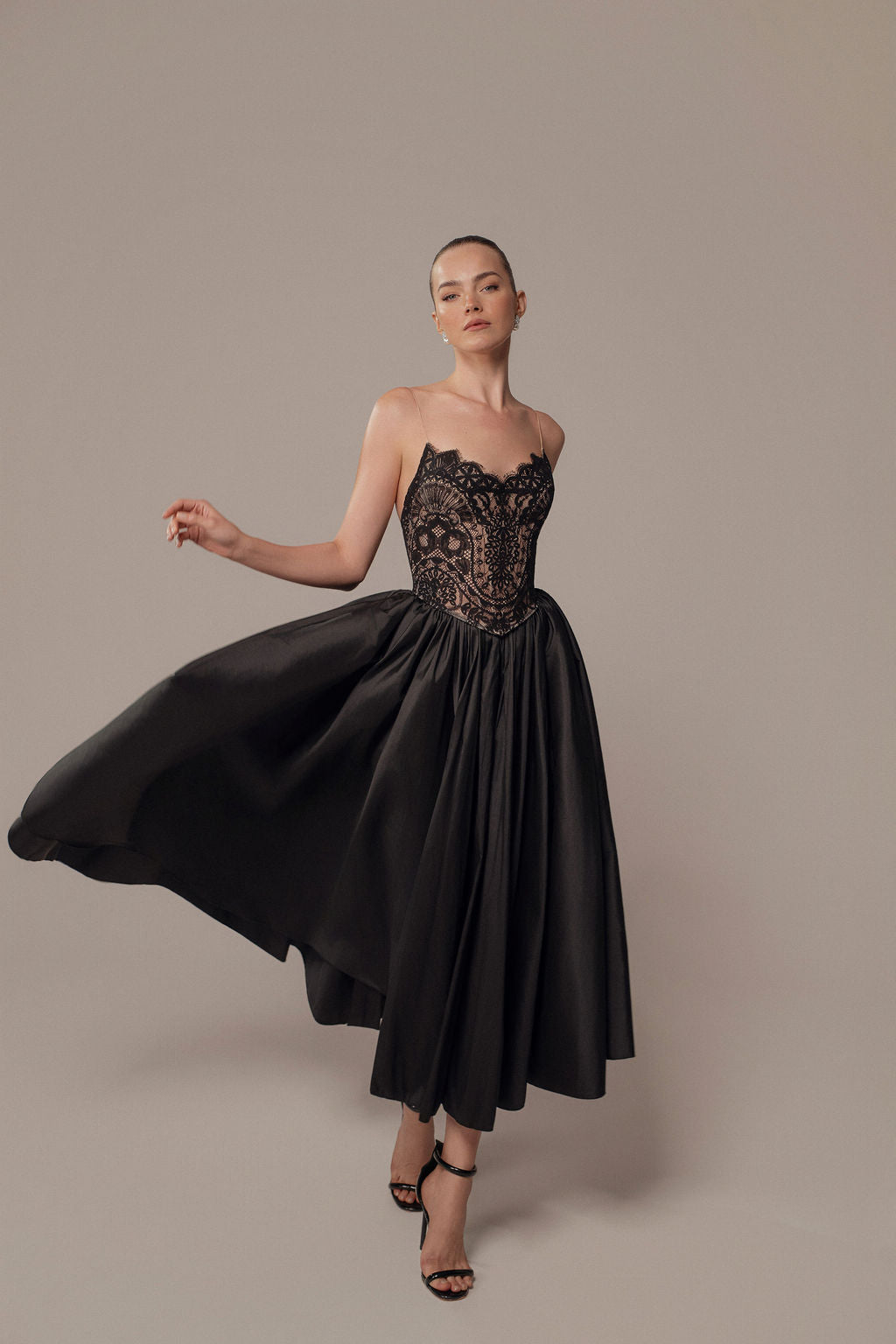 אישה מצטלמת באלגנטיות על רקע חלק בשמלת Luna מקולקציית הערב 2026 של דרור קונטנטו, עם תחרה שחורה ללא כתפיות וחצאית מלאה, בשילוב עם סנדלים שחורים עם עקבים גבוהים.