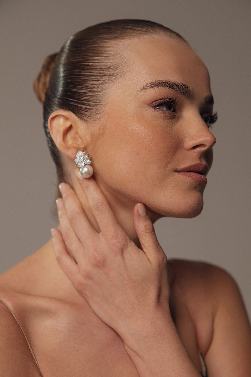 אישה עם שיער חלק ומסודר בקוקו נוגעת בעדינות בצווארה, ומציגה את העגילים האלגנטיים Gia מקולקציית Evening 2026. היא מאופרת באיפור טבעי, גבות חומות רכות, ומראה רגוע ובטוח.