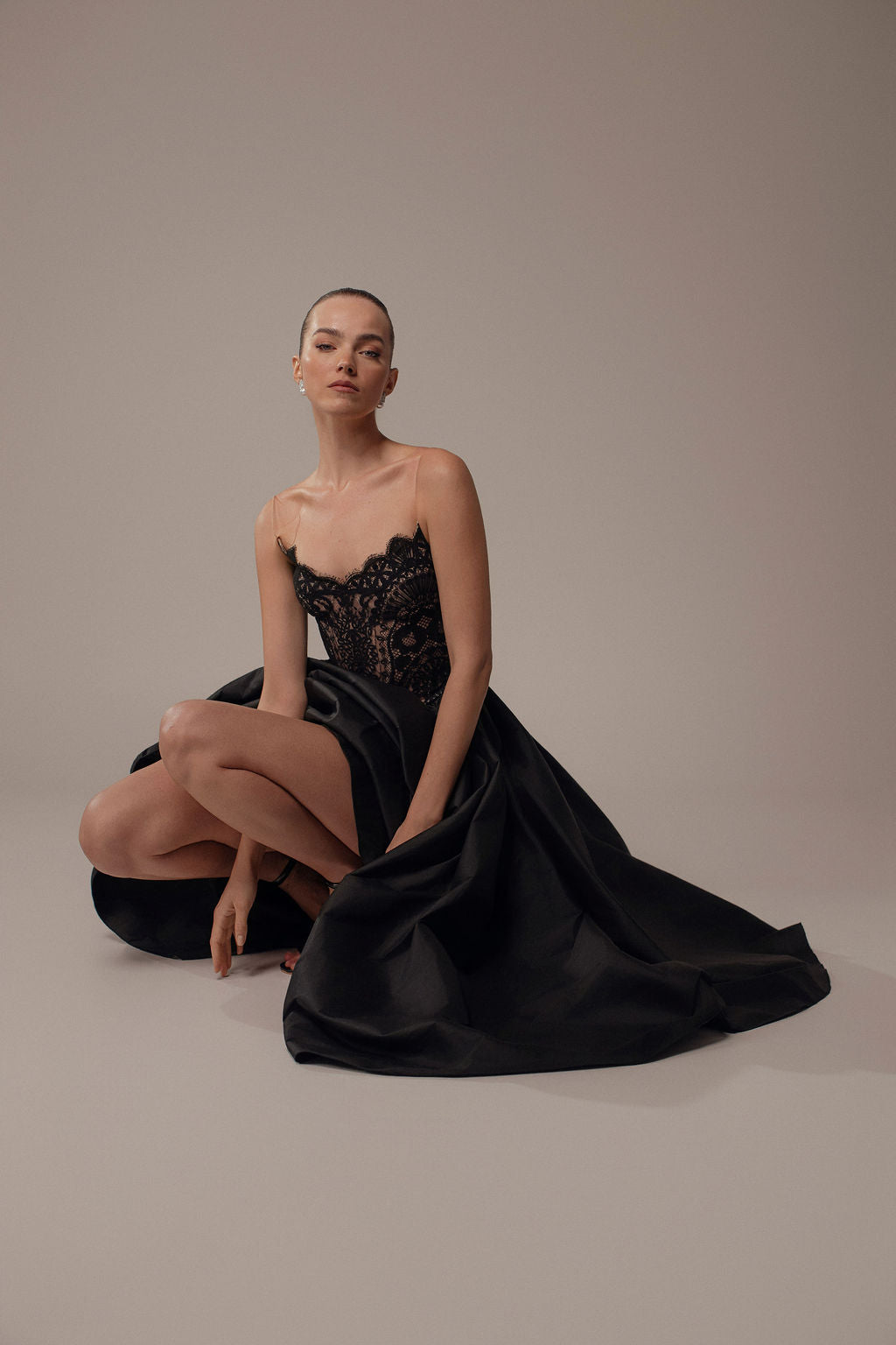 אישה לבושה בשמלת Luna מקולקציית Evening 2026 יושבת על הרצפה, ברך אחת מורמת, ומביטה בביטחון למצלמה על רקע ניטרלי.