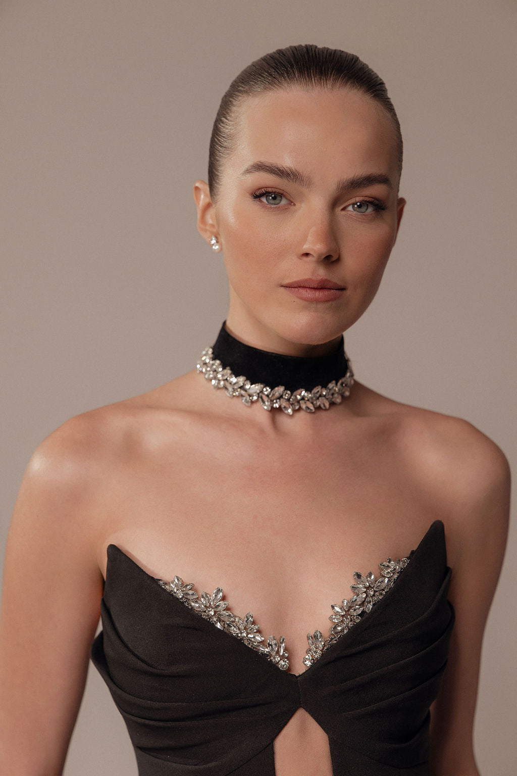 אישה עם שיער משוח לאחור לובשת את שמלת Maya מקולקציית ערב 2026 — שמלה שחורה ללא כתפיות, עם עיטורים כסופים ושרשרת שחורה משובצת אבנים — ומצטלמת על רקע בז' חלק.