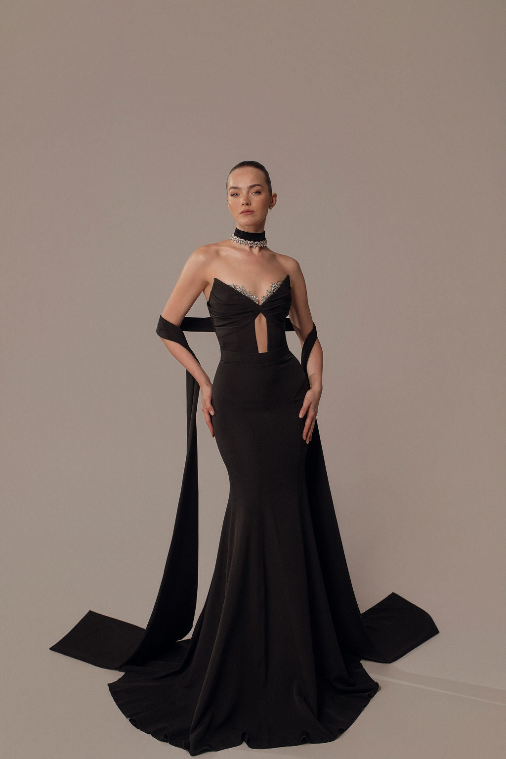 אישה מדגמנת את שמלת Maya מקולקציית הערב 2026 של דרור קונטנטו: שמלת ערב צמודה באורך הרצפה עם מחשוף לב, חיתוך מפתח, אבנטים דרמטיים על הזרועות ושרשרת צוואר נועזת — על רקע חלק.