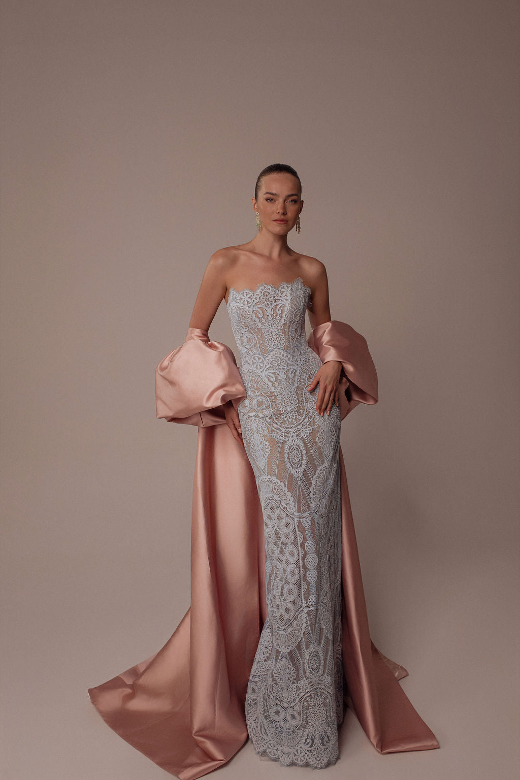 אישה מצטלמת על רקע אחיד בשמלת Elia מקולקציית הערב 2026 של דרור קונטנטו, שמלה ללא כתפיות, צמודה לגוף, עם תחרה בעלת דוגמאות מורכבות ושובל סאטן ורוד בהיר. היא משלימה את המראה עם תכשיטים מינימליים.