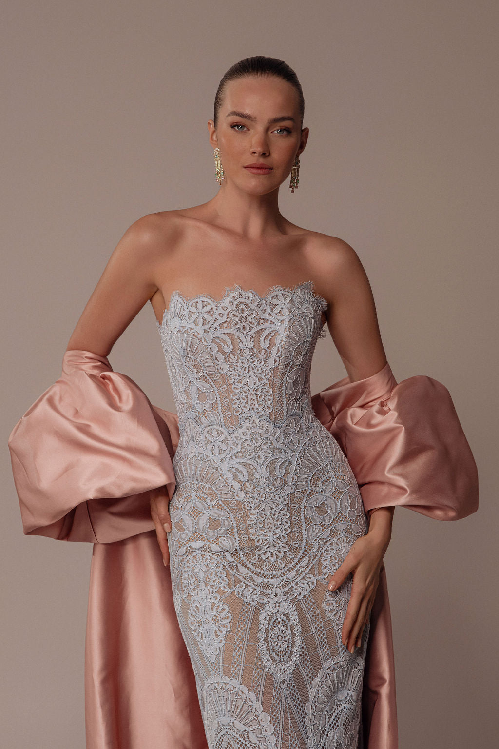 אישה מדגמנת את שמלת אליה ללא כתפיות, עם תחרה בעלת דוגמה מורכבת, מקולקציית הערב 2026 של דרור קונטנטו, עטופה בצעיף משי ורוד בהיר, שיערה אסוף לאחור ועגילי סטטמנט מדגישים את מראה האלגנטי שלה.