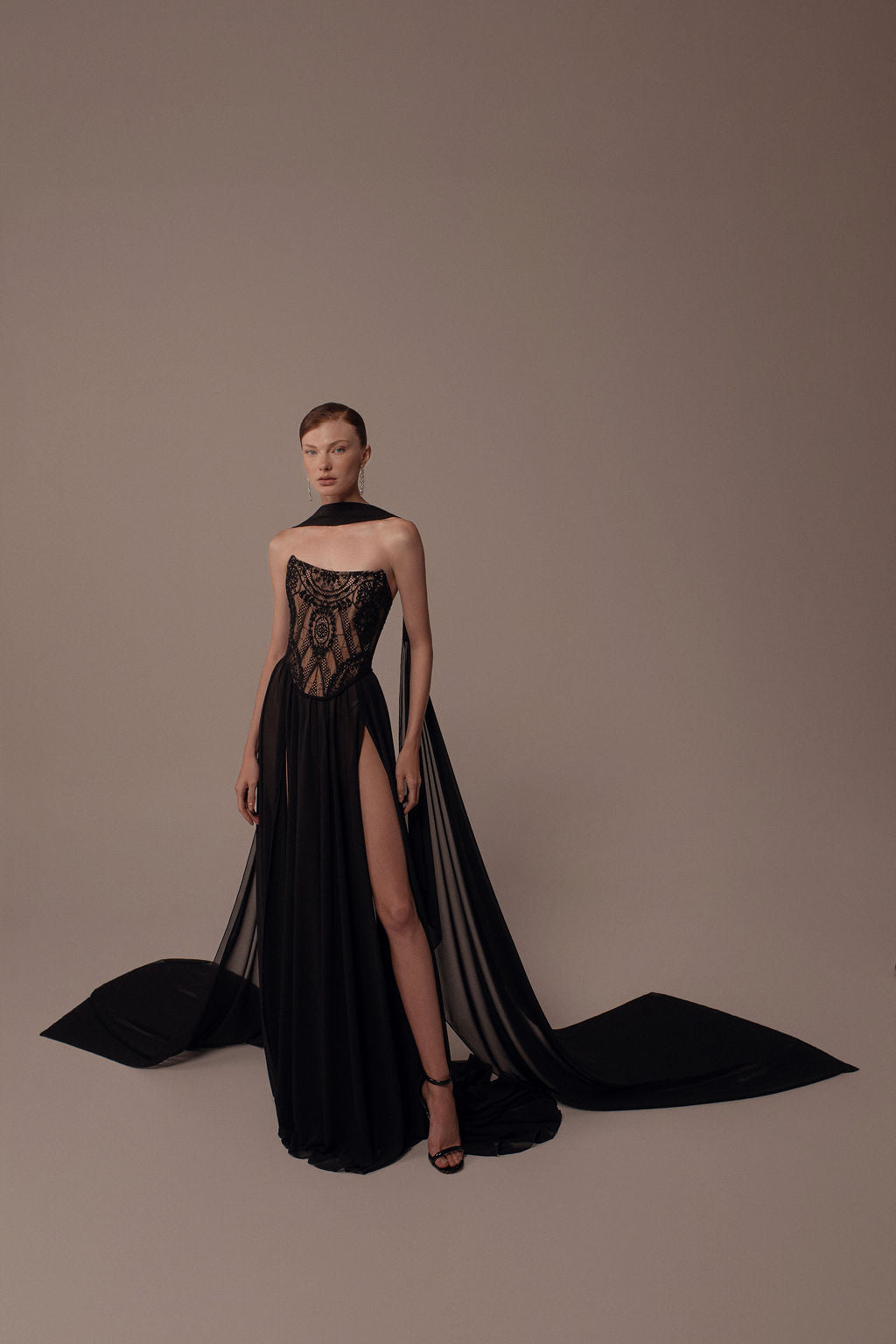 אישה מצטלמת בסטודיו כשהיא לובשת את שמלת Alma מקולקציית הערב 2026 של דרור קונטנטו — שמלת ערב שחורה ללא כתפיות עם מחוך תחרה שקוף, חתך גבוה ושובל מתנפנף. שיערה אסוף לאחור והיא נועלת נעלי עקב שחורות על רקע חלק.