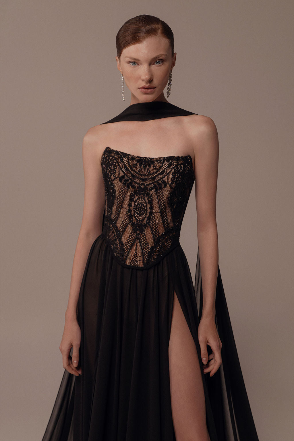 אישה עומדת על רקע בז' חלק בשמלת ערב שחורה ללא כתפיות מדגם "אלמה" מקולקציית הערב 2026 של דרור קונטנטו, עם מחשוף תחרה מורכב, חצאית עם חתך גבוה וצעיף שחור שקוף כרוך סביב צווארה.