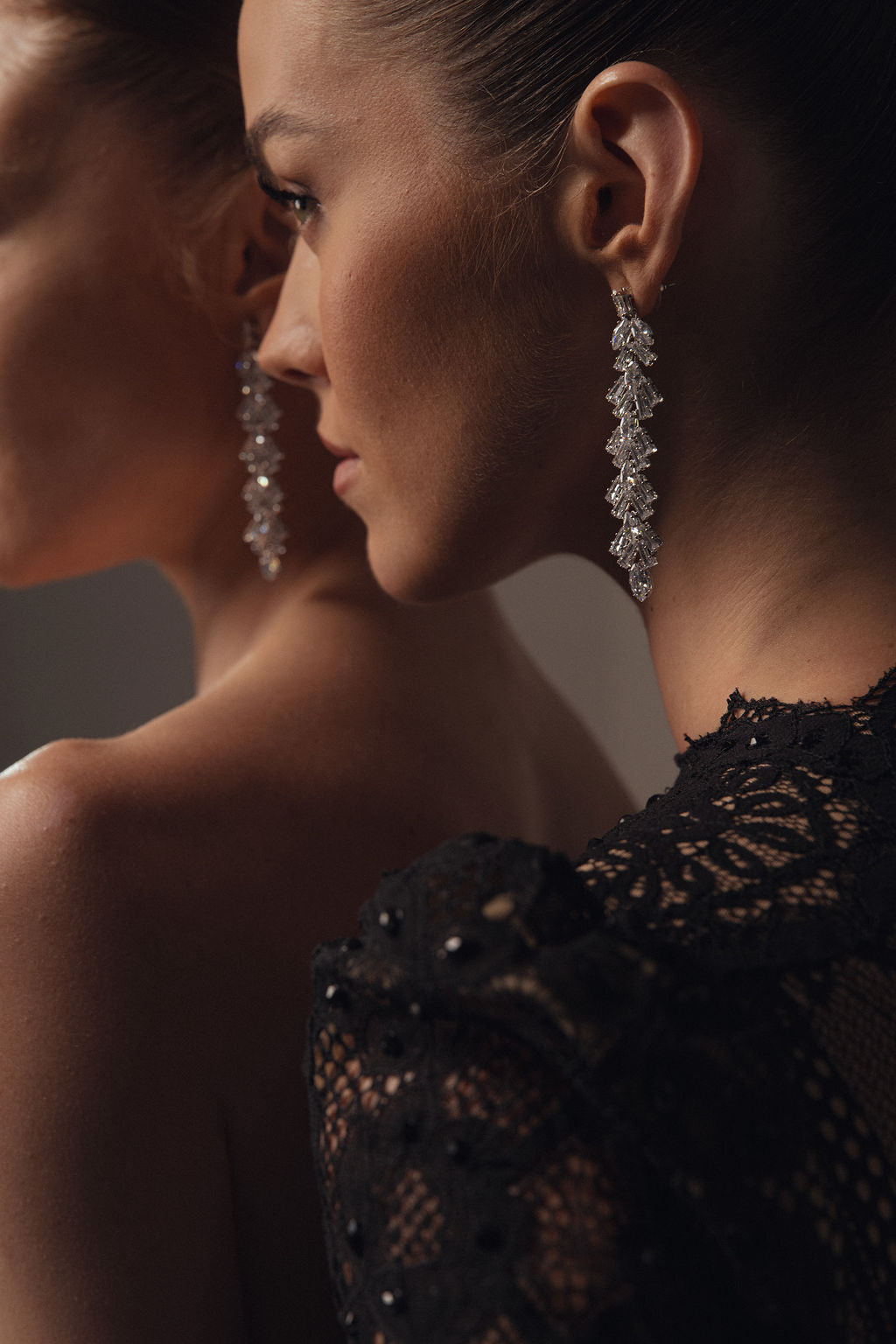 שתי נשים בפרופיל עונדות את עגילי Vera מקולקציית Evening 2026, בשילוב עם תלבושות תחרה שחורות ואלגנטיות. עיצובים נוצצים בסגנון נברשת ופרטי תחרה מודגשים על ידי התאורה.