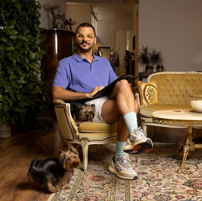 גבר המרכיב משקפיים, לבוש בחולצת פולו כחולה ומכנסיים קצרים, יושב על כיסא וינטג' עם מחשב נייד על ברכיו. שני כלבים קטנים יושבים לידו בסלון נעים ומקושט, עם צמחים ושטיחים מעוצבים.