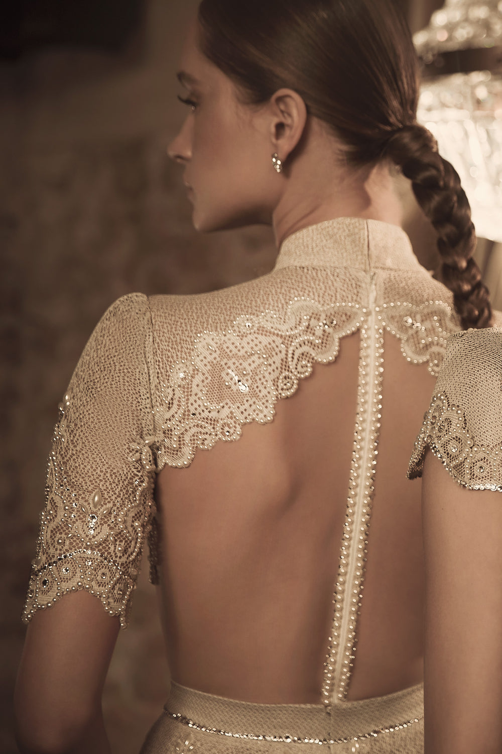 אישה עם צמה לובשת את שמלת הכלה Blaire + Margo מקולקציית הכלות 2018 של דרור קונטנטו, עם תחרה, פאייטים, שרוולים קצרים, גב פתוח, צווארון גבוה וכפתורי פנינה, מוצגת מאחור בתאורה חמה ורכה.