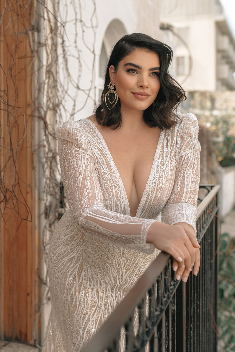 אישה עם שיער כהה לובשת את שמלת הכלה הלבנה והנוצצת "אליסון" מקולקציית Bridal Plus 2022 של דרור קונטנטו, נשענת על מעקה שחור בחוץ ומחייכת ברכות. היא משלימה את מראה שמלות כלה שלה עם עגילי זהב גיאומטריים גדולים.