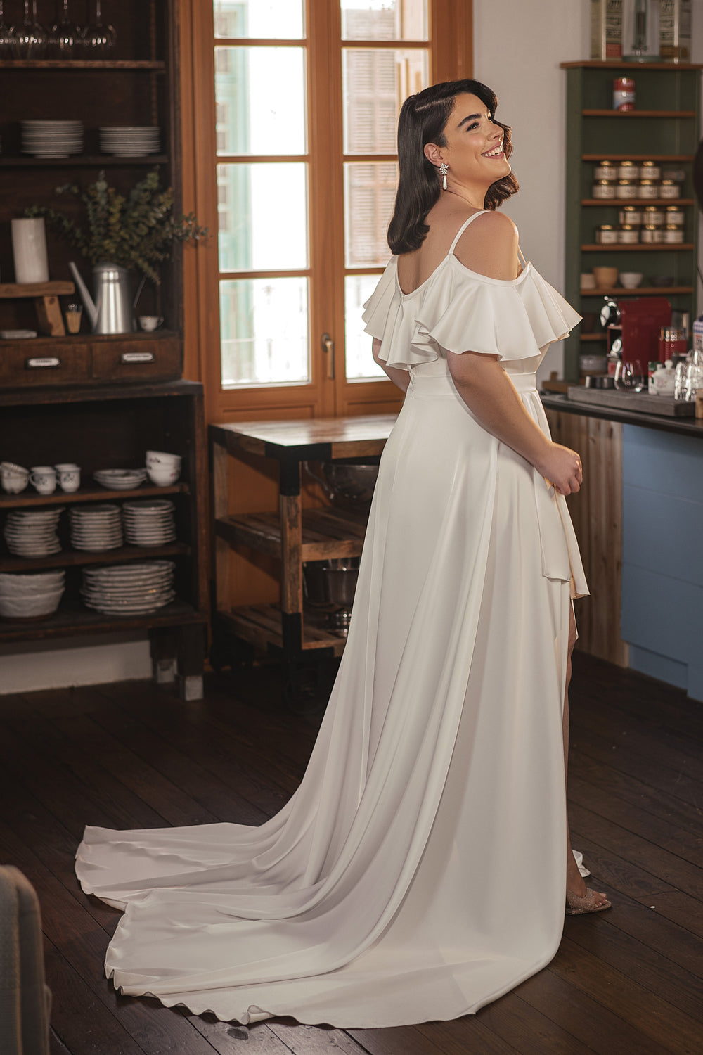 אישה מקולקציית Bridal Plus 2022 - שמלת כלה לבנה עם כתפיים חשופות של Nala מחייכת במטבח נעים עם ארונות עץ, מדפי כלים ואור שמש הזורם מבעד לחלונות.