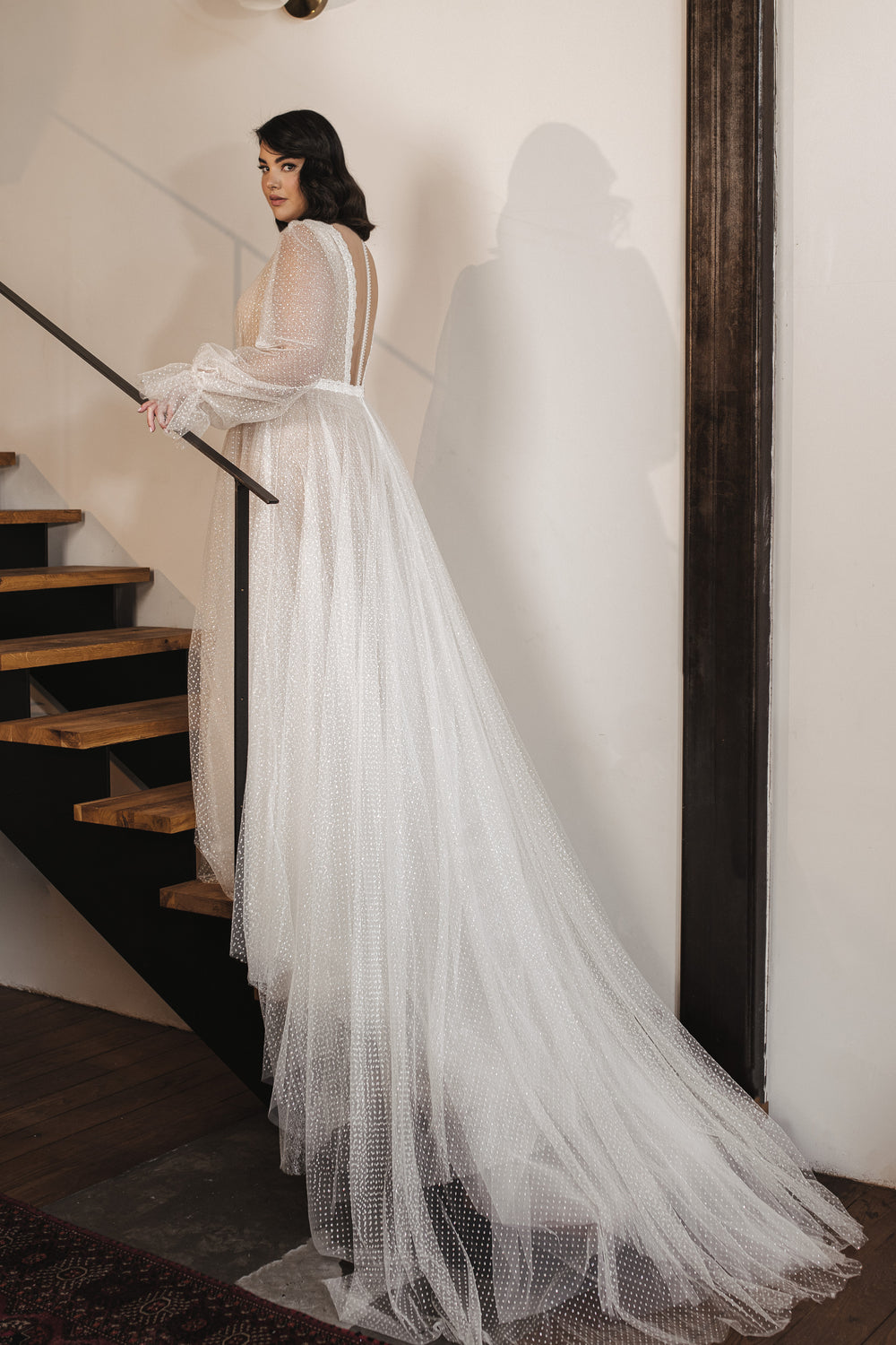 אישה לובשת את שמלת Livia מקולקציית Dror Kontento Bridal Plus 2022 — שמלה ארוכה, שקופה, עם נקודות לבנות — בעודה עומדת על גרם מדרגות עץ בחלל מודרני ומואר באור רך.