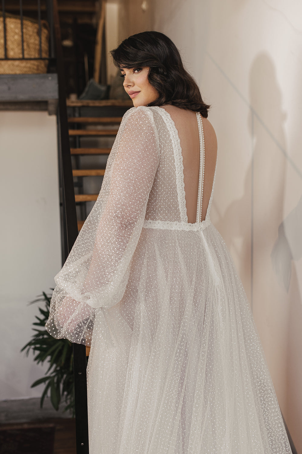 אישה עם שיער כהה לובשת את שמלת הכלה Livia השקופה עם שרוולים ארוכים מקולקציית Bridal Plus 2022 של דרור קונטנטו, עם נקודות פולקה מרקמות, כשהיא מצטלמת בתוך הבית ליד גרם מדרגות, ומציגה את העיצוב האלגנטי של שמלות כלה.