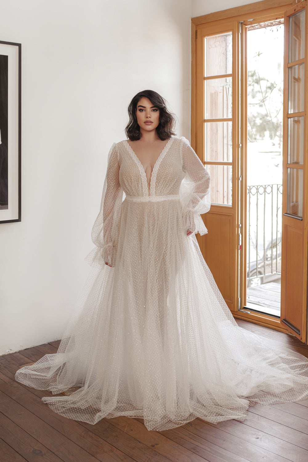 אישה עומדת בתוך הבית ליד דלת עץ פתוחה, לבושה בשמלת כלה מקולקציית Bridal Plus 2022 - Livia. העיצוב כולל מחשוף V עמוק, שרוולים ארוכים שקופים וחצאית טול זורמת עם נקודות.