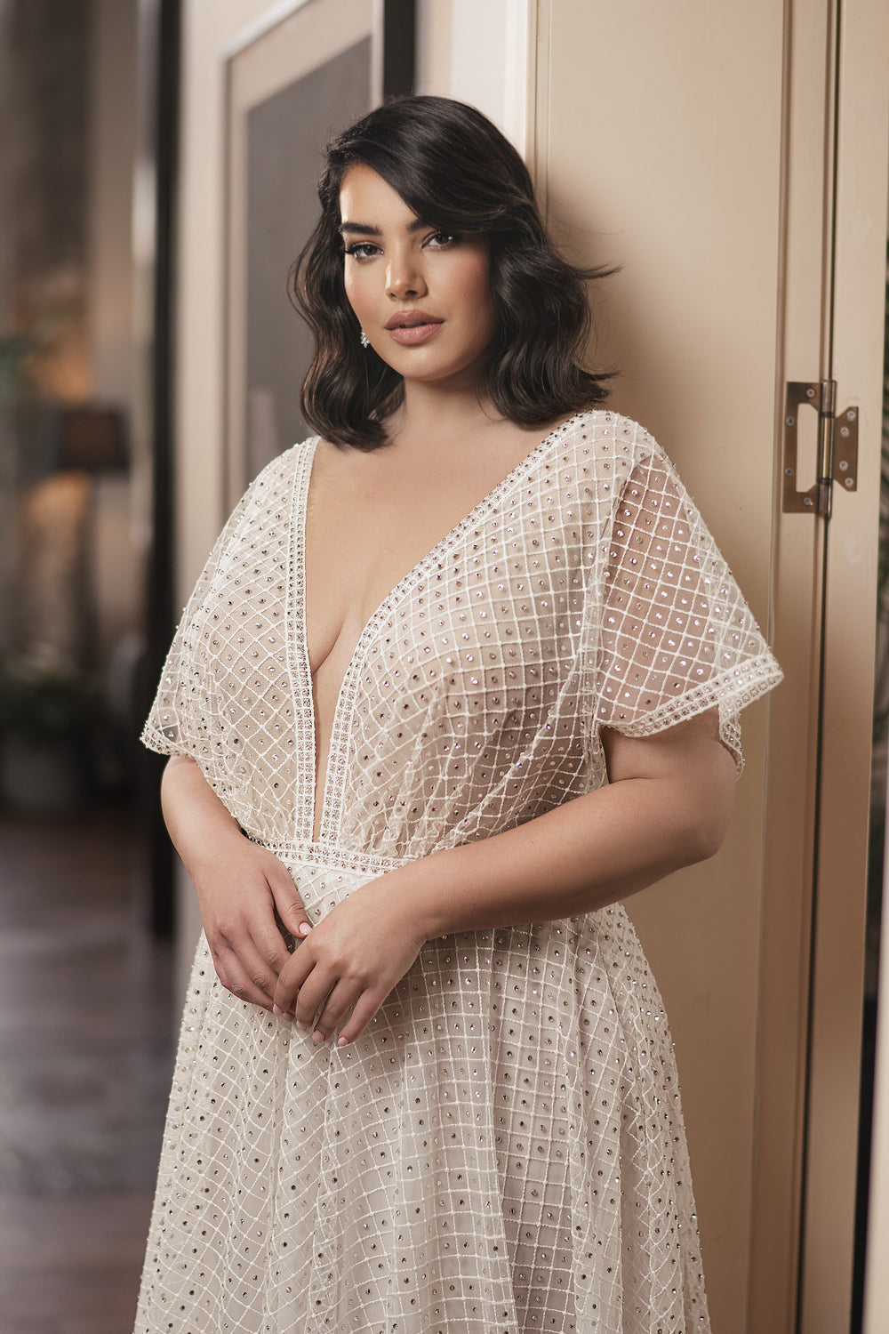 אישה עם שיער כהה וגלי לובשת את קולקציית Bridal Plus 2022 - Jolie, שמלת כלה לבנה עם מחשוף V עמוק ומחוטבת בחרוזים, בעיצוב דרור קונטנטו. היא עומדת בתוך הבית, ידיה שלובות, ומביטה במבט רך למצלמה על רקע מטושטש.