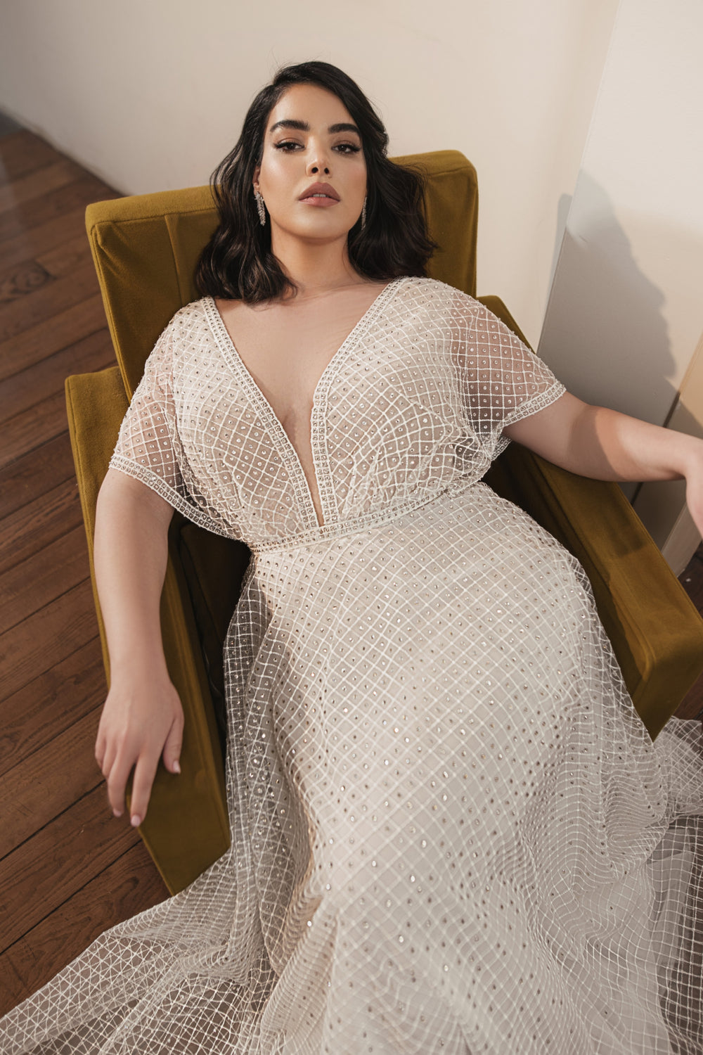 אישה עם שיער כהה באורך הכתפיים נשענת על כיסא בצבע צהוב חרדל, לבושה בשמלת הכלה Jolie מקולקציית Bridal Plus 2022. ברקע ניתן לראות רצפת עץ וקיר בהיר.