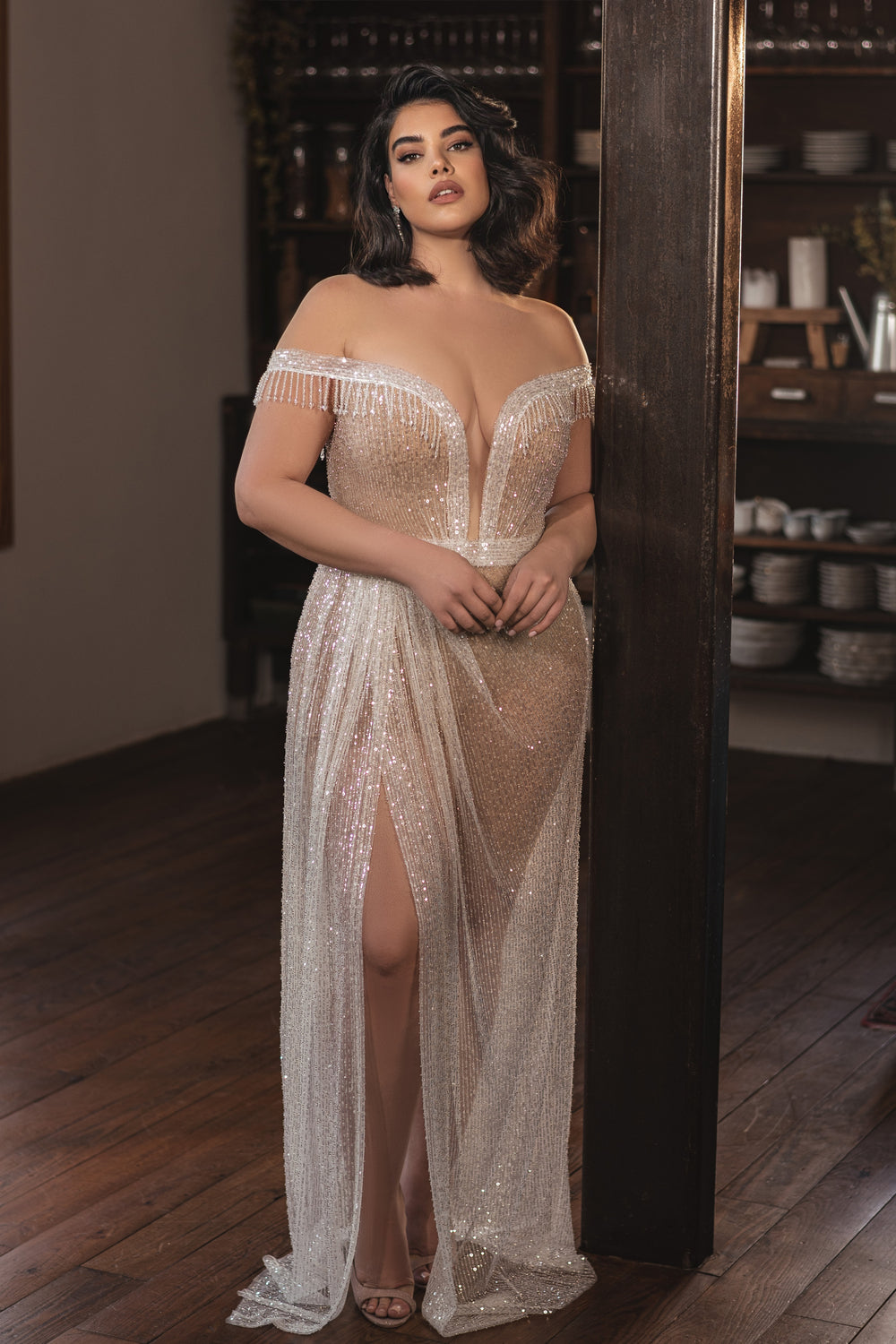 אישה לבושה בשמלת "Bridal Plus Collection 2022 - Estelle" מנצנצת, עם כתפיים חשופות ואורך עד הרצפה, עומדת בתוך הבית על רצפת עץ, נשענת על קורה עם מדפי כלים ברקע.