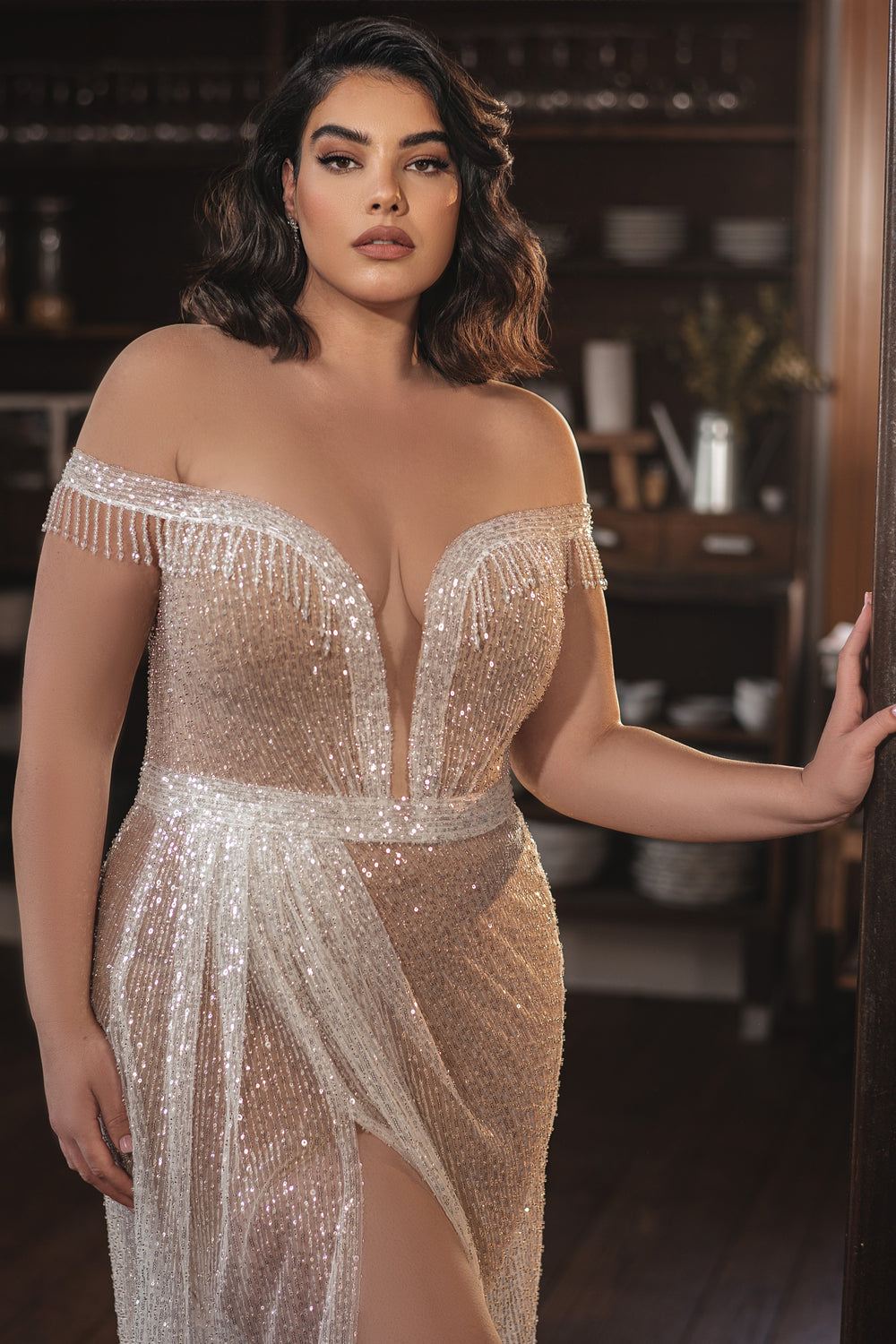 אישה מקולקציית Bridal Plus 2022 - Estelle, שמלת חתונה מנצנצת עם כתפיים חשופות ומחוטבת בחרוזים, עומדת בחינניות באור חם, ומדגימה את האלגנטיות והסגנון של שמלות כלה במידות גדולות.