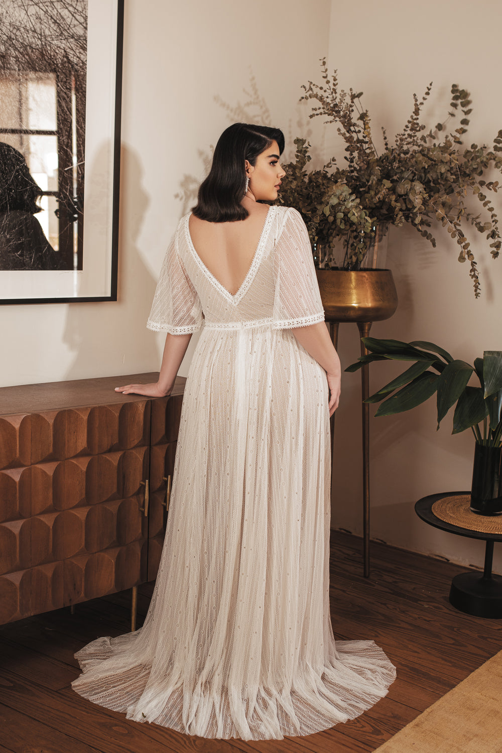 אישה לבושה בשמלת כלה לבנה וזורמת מקולקציית Bridal Plus 2022 - Jenesis של דרור קונטנטו עומדת בתוך הבית, עם גבה למצלמה, יד אחת מונחת על ארון עץ, מוקפת בצמחים ירוקים ובתפאורה בגוונים ניטרליים.