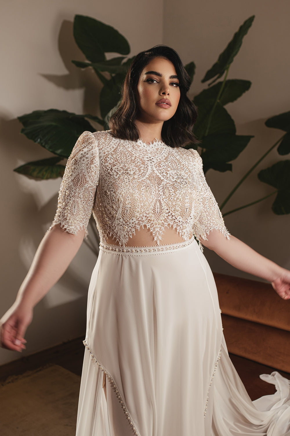 אישה מקולקציית Bridal Plus 2022 - שמלת Pearl, עם תחרת תכשיטים אלגנטית וחצאית שיפון זורמת, עומדת בתוך הבית ליד צמח ירוק גדול, ומביטה בביטחון למצלמה.