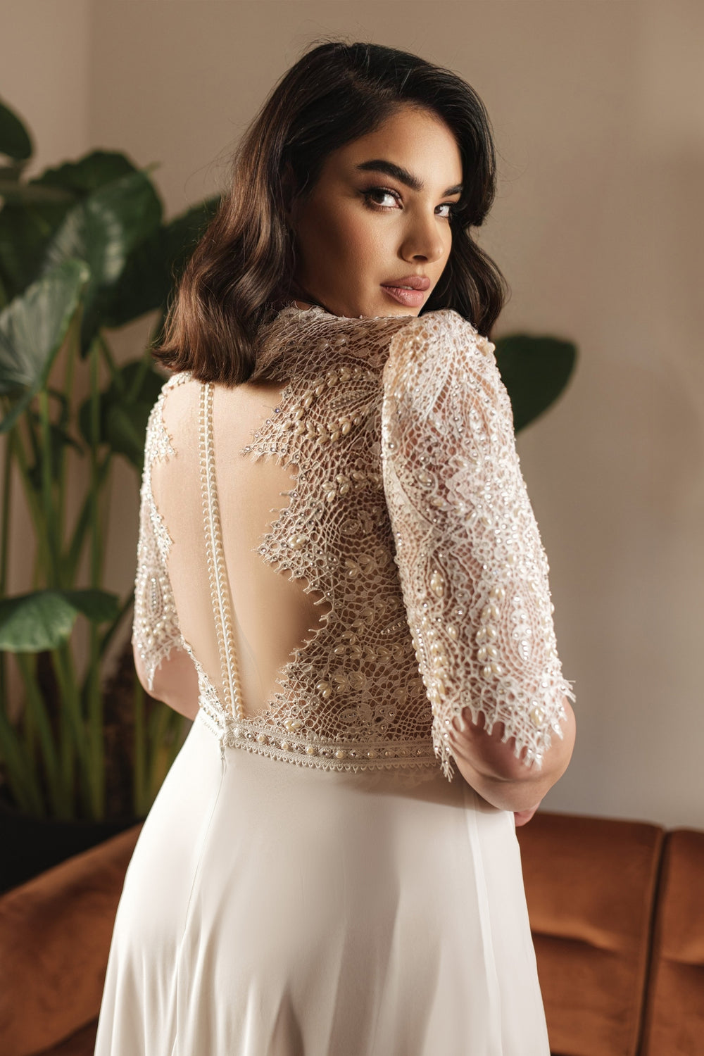 אישה עם שיער כהה וגלי מדגמנת את קולקציית Bridal Plus 2022 - שמלת Pearl, שמלה לבנה עם חצאית שיפון ותחרת תכשיטים בעבודת יד על הגב והשרוולים, עומדת בתוך הבית ליד צמחים וספה חומה.