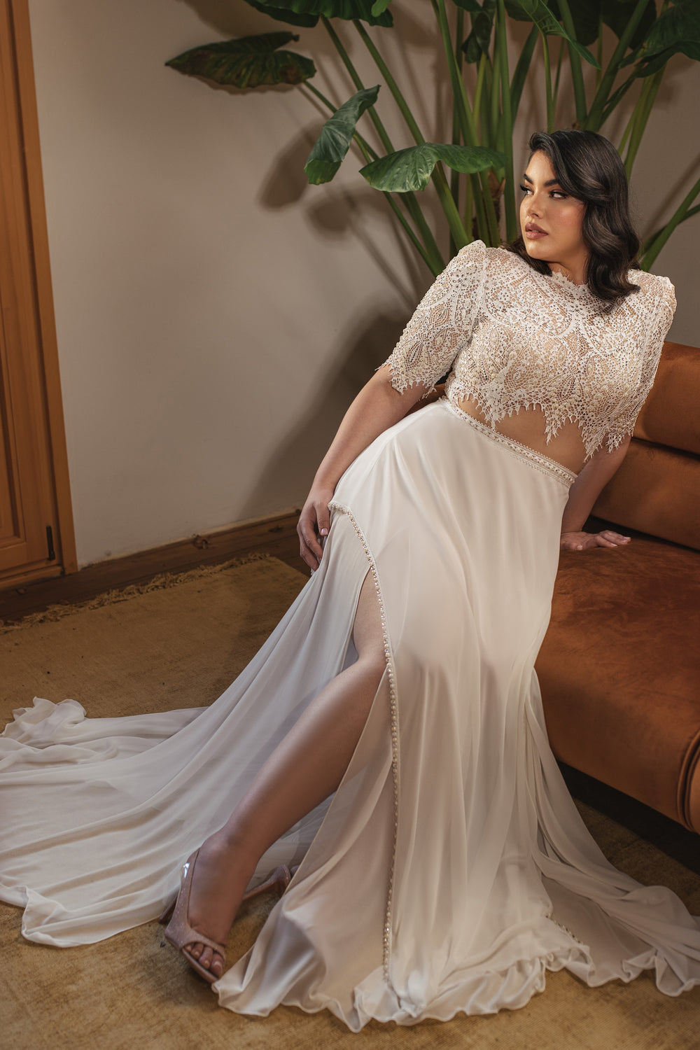 אישה מקולקציית Bridal Plus 2022 - Pearl, לבושה בחולצת בטן קצרה מעוטרת בתכשיטים בעבודת יד וחצאית שיפון עם חתך גבוה, פוזלת באלגנטיות על ספה חומה עם צמח ירוק ולוחות עץ ברקע.