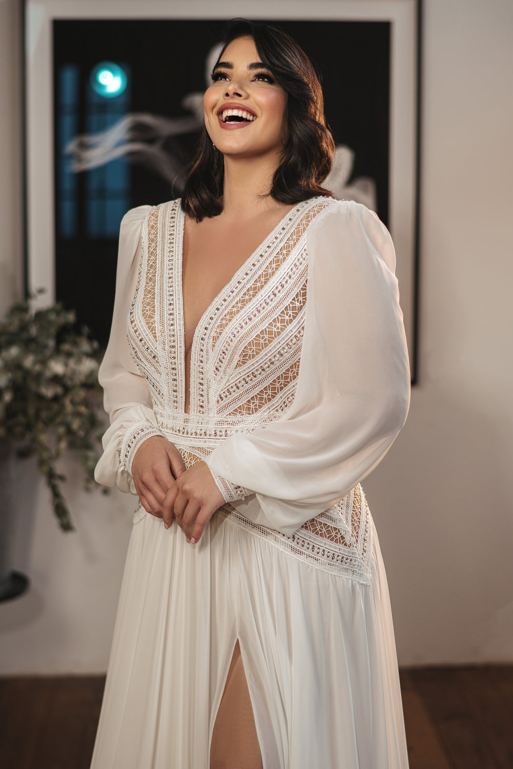 אישה מחייכת בתוך הבית בקולקציית Bridal Plus 2022 - Bellamy: שמלה לבנה עם שרוולים ארוכים, מחוך תחרה משובץ אבנים, מחשוף V עמוק, חתך גבוה וחצאית שיפון. צמחים מטושטשים ואמנות ממוסגרת מדגישים את הרקע.