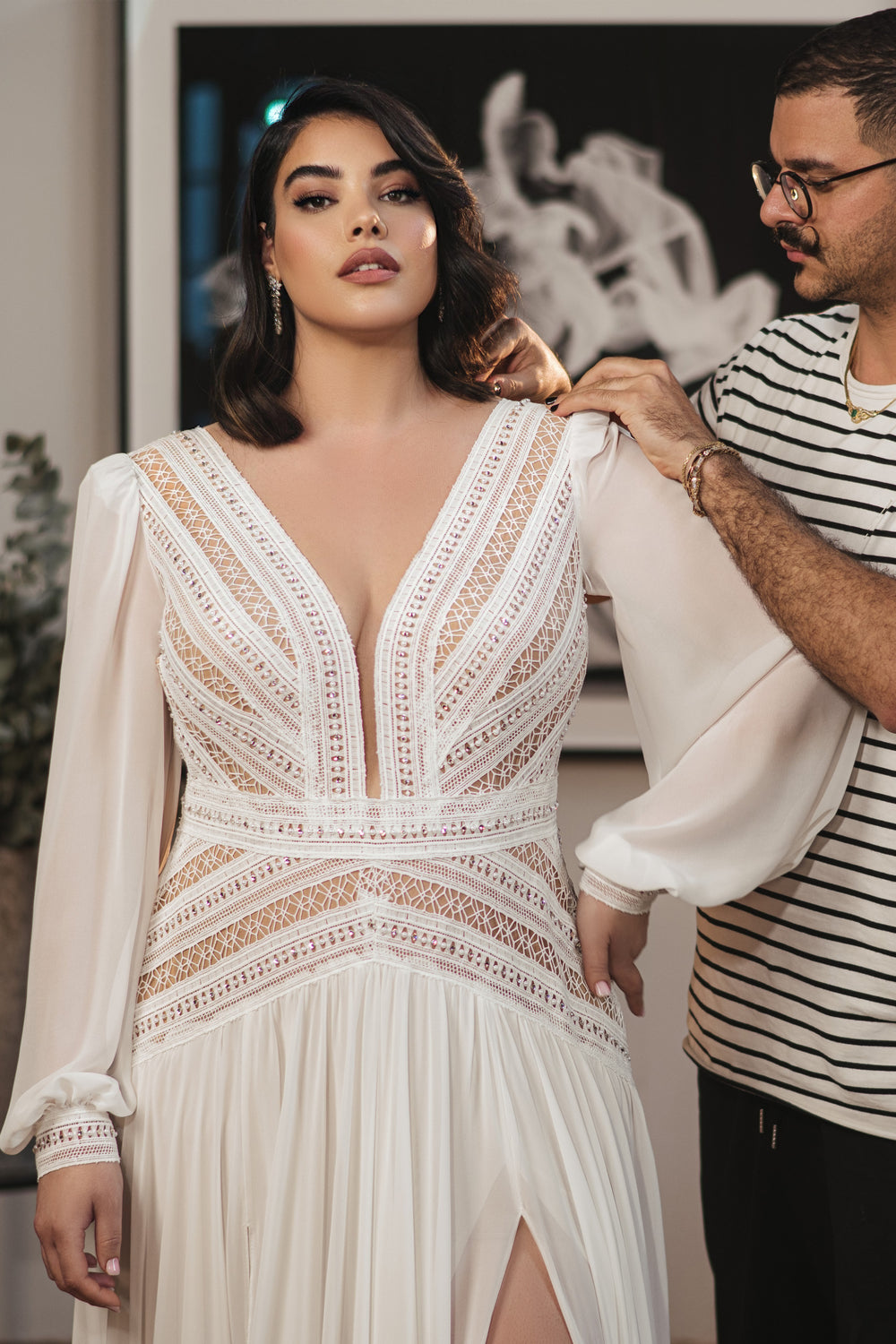 אישה לובשת את קולקציית Bridal Plus 2022 - Bellamy, שמלה לבנה עם שרוולים ארוכים וחלק עליון מתחרה מעוטר באבני חן, בזמן שמישהו עם משקפיים וחולצה מפוספסת מסדר את תלבושתה. ברקע נראית יצירת אמנות מטושטשת בשחור-לבן.