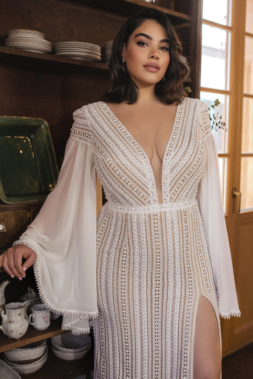 אישה עם שיער כהה באורך הכתפיים לובשת את שמלת "Bridal Plus Collection 2022 - Karla", שמלת תחרה בעבודת יד עם שרוולים מתרחבים שקופים ומחשוף V עמוק, עומדת בתוך הבית ליד מדפי כלים ומביטה בביטחון למצלמה.