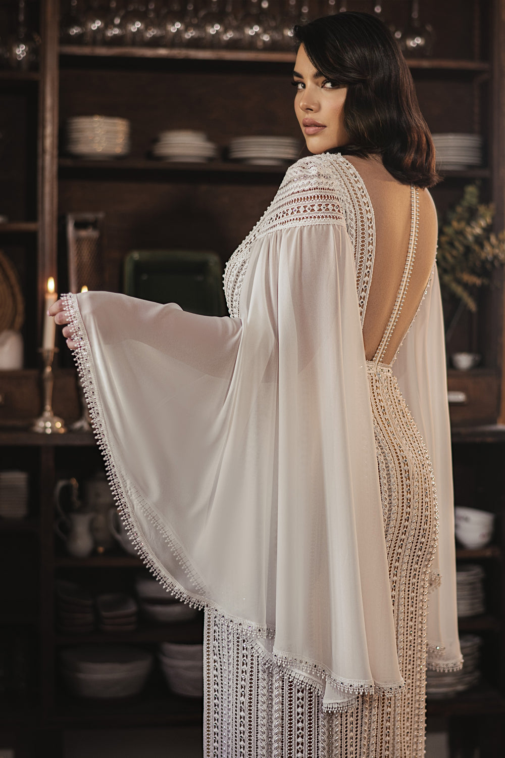 אישה לובשת את קולקציית Bridal Plus 2022 - Karla, שמלת תחרה מעוטרת באבני חן בעבודת יד של דרור קונטנטו. בתוך הבית, היא מושיטה שרוול רחב; ברקע נראים מדפים עם צלחות וכלי מטבח, המדגישים את השמלה המדהימה.