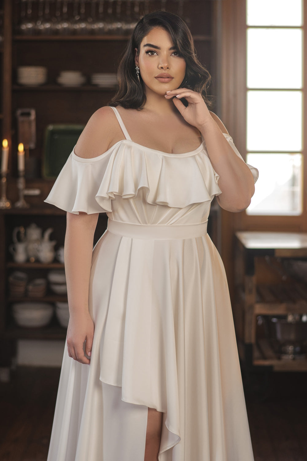 אישה לבושה בשמלת Nala מקולקציית Bridal Plus 2022 עומדת בתוך הבית, יד אחת על פניה, עם שיער ארוך וכהה, מדפי כלים וחלונות מוארים באור השמש ברקע.