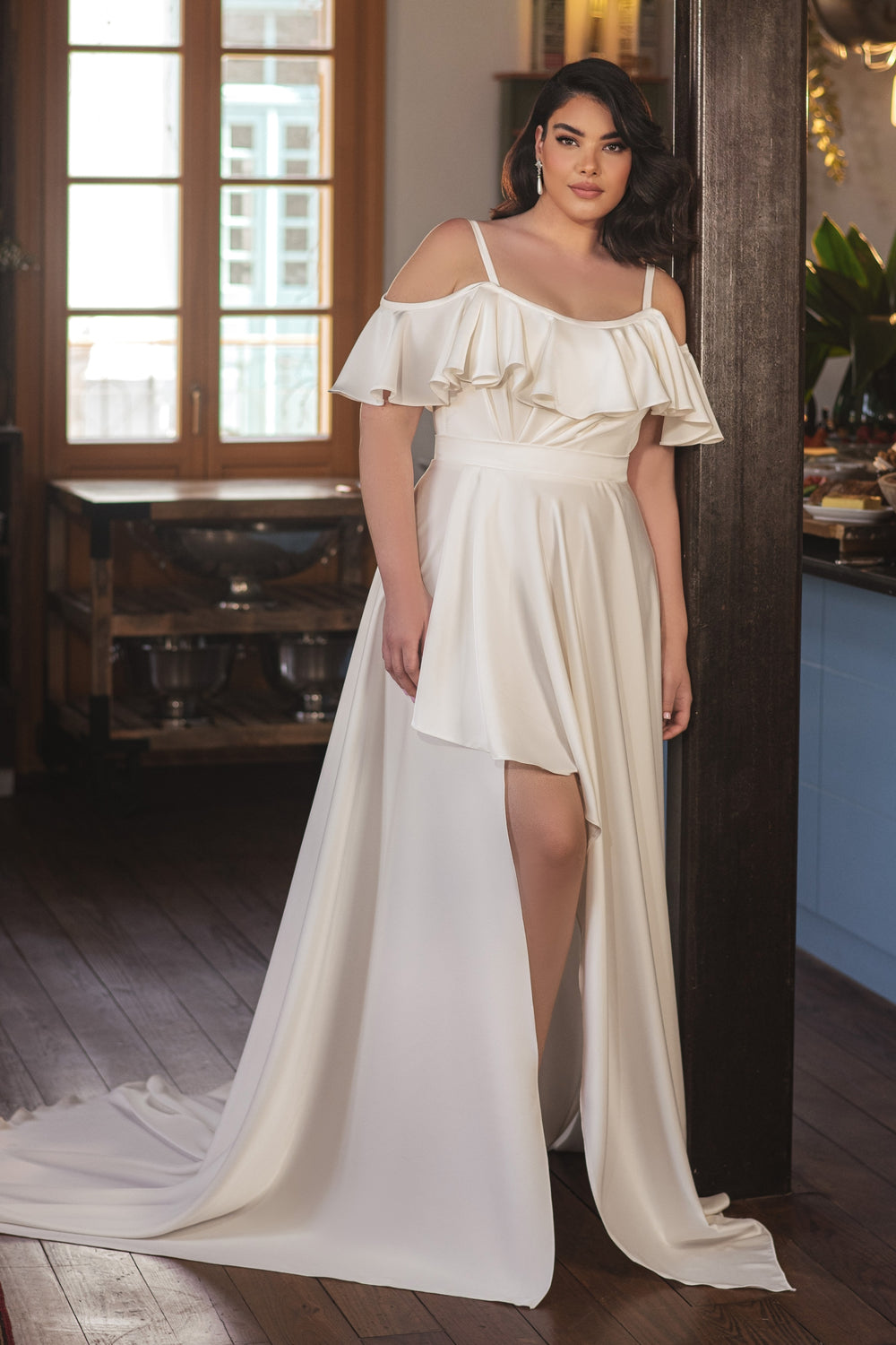 אישה לבושה בשמלת כלה לבנה עם כתפיים חשופות מקולקציית Bridal Plus 2022 - Nala עומדת בתוך הבית ליד עמוד עץ, כשמאחוריה אור טבעי זורם מבעד לחלון גדול.