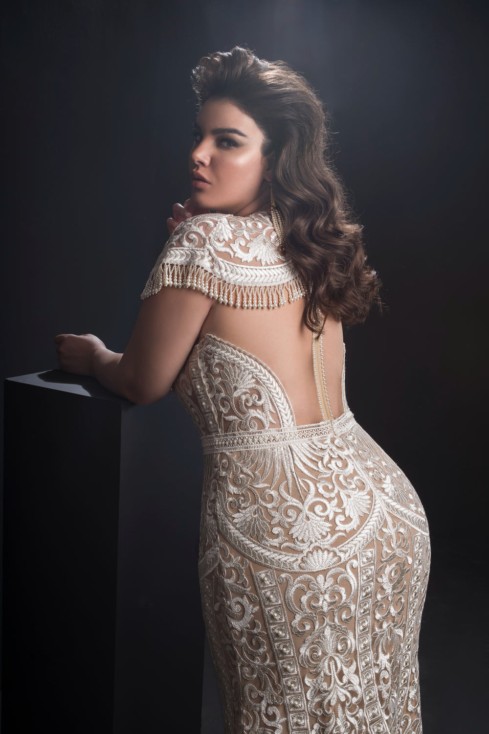 אישה עם שיער ארוך וגלי מצטלמת בשמלת אמלי המעוטרת והחשופה מגב, מקולקציית Bridal Plus 2019, עם תחרה מורכבת ופרטים של שוליים, על רקע כהה עם תאורה דרמטית.