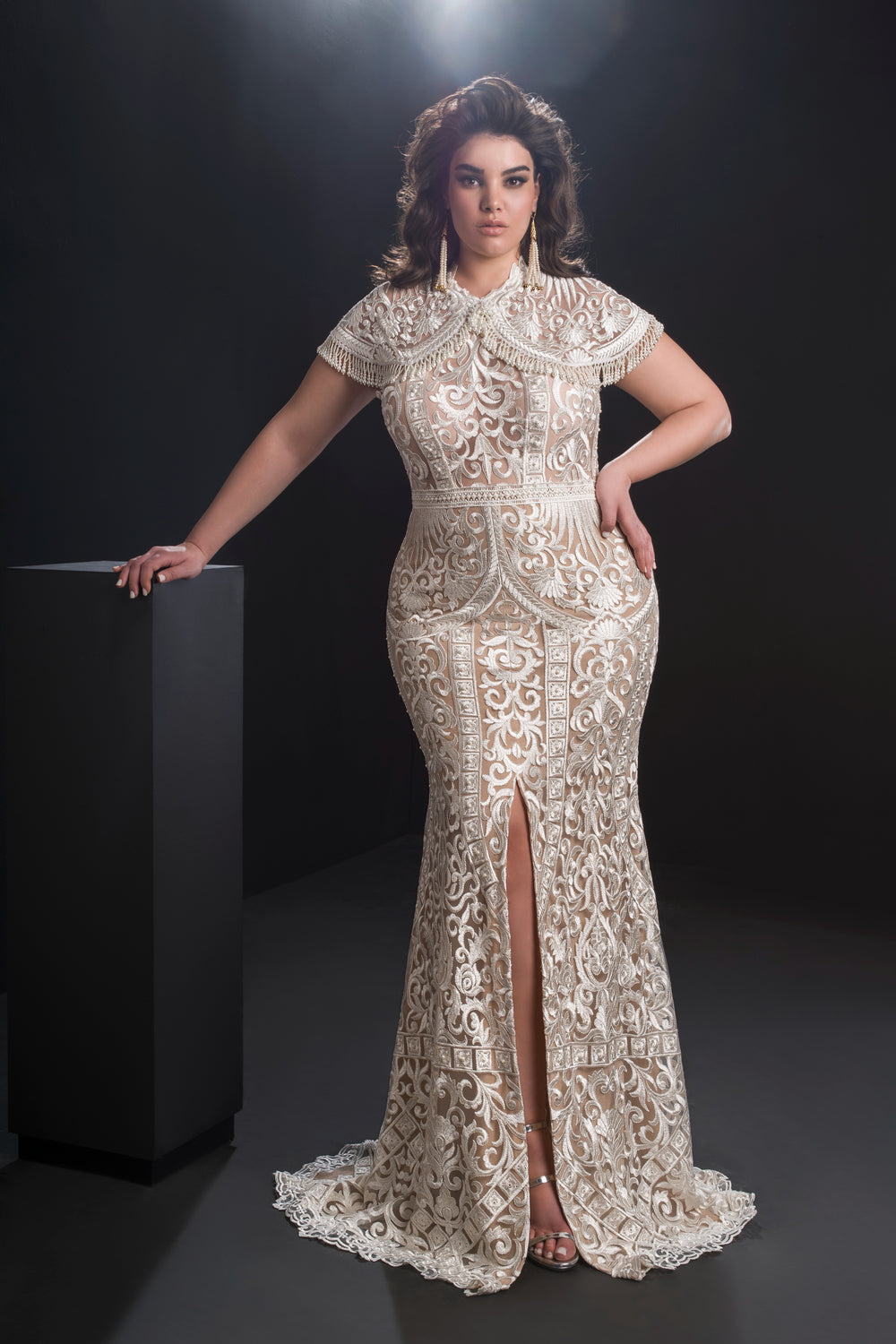 אישה לבושה בשמלת "אמלי" אלגנטית, מחויטת וארוכה עד הרצפה מקולקציית Bridal Plus 2019 עומדת בביטחון על רקע כהה, כשהיא מניחה יד אחת על כן שחור.