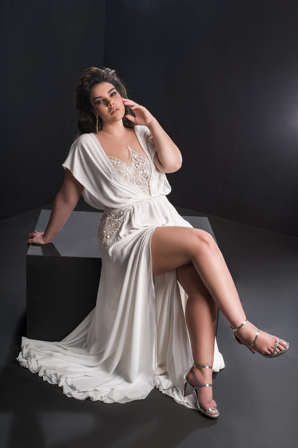 אישה בשמלת כלה זורמת מקולקציית Bridal Plus 2019 - Adele יושבת על פלטפורמה שחורה, שיערה הגלי והכהה מסודר באלגנטיות. היא נועלת סנדלים כסופים ומצטלמת בחינניות על רקע כהה.