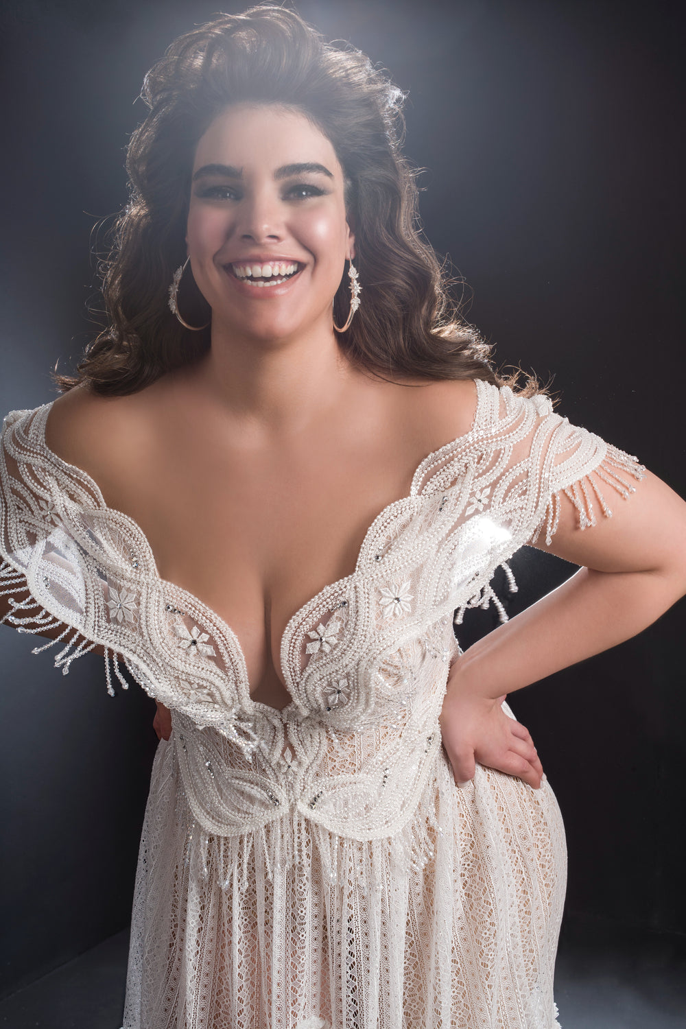 אישה עם שיער ארוך, גלי וכהה מחייכת חיוך רחב, לבושה בשמלת Giovanna מקולקציית Bridal Plus 2019, בעיצוב עם כתפיים חשופות, רקמה מורכבת, מחשוף עמוק ופרטים עם שוליים, ומצטלמת בביטחון על רקע כהה.