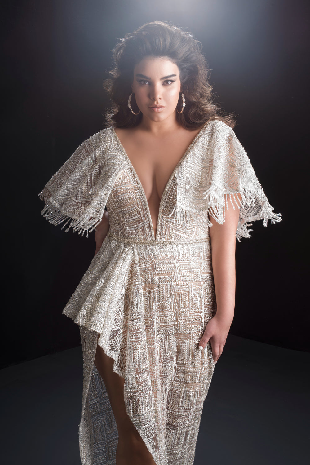 אישה עם שיער גלי ושופע מצטלמת בשמלת הכלה הנוצצת Bridal Plus Collection 2019 - Paula, עם מחשוף V עמוק ושרוולים רחבים עם שוליים, על רקע כהה המואר באור רך וזוהר.