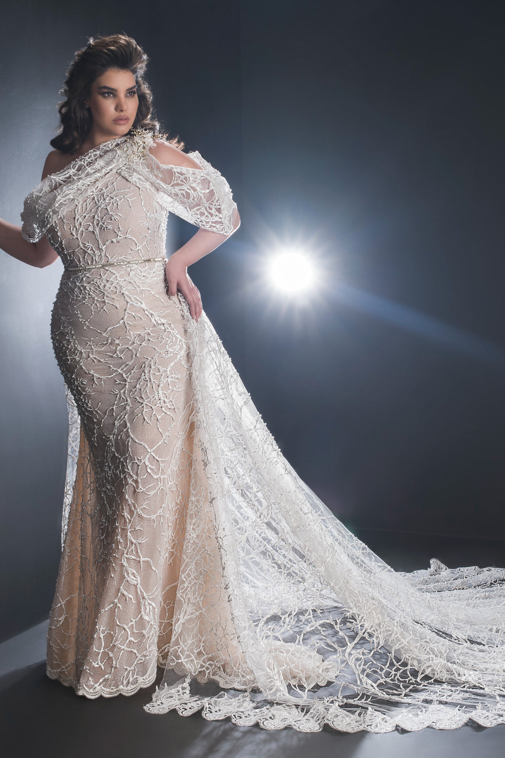 אישה מדגמנת את קולקציית Bridal Plus 2019 - שמלת Royal של Dror Kontento, עם עיצוב תחרה אלגנטי עם כתפיים חשופות, בדוגמת פרחים לבנים על רקע בצבע גוף, המודגשת על ידי תאורה דרמטית ליצירת אפקט זוהר.