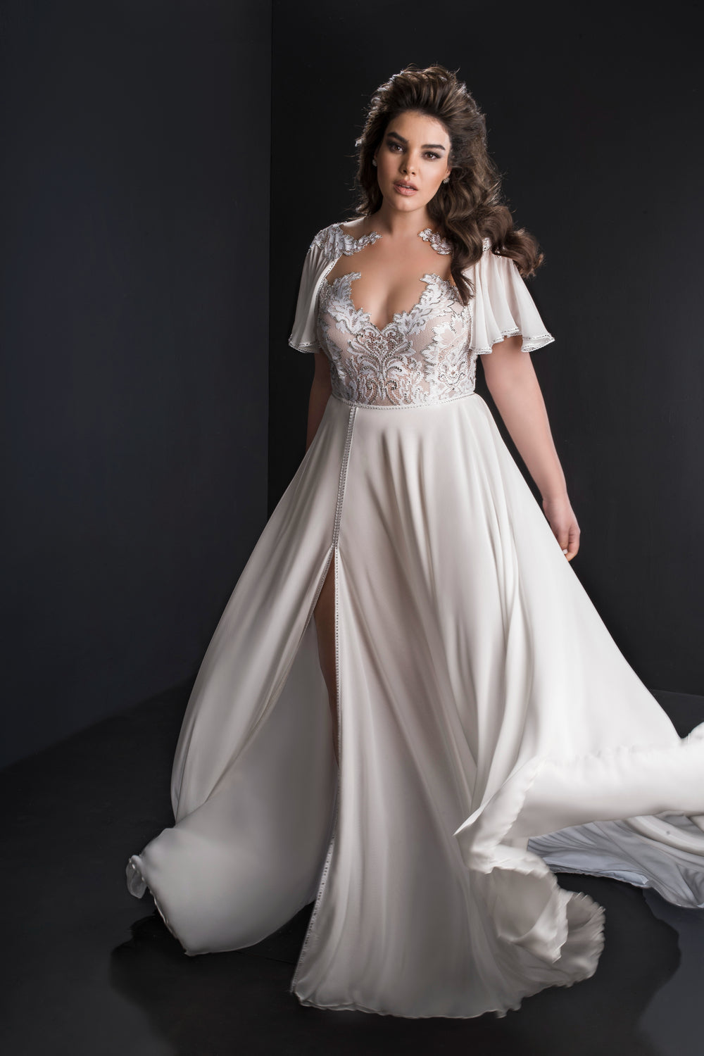 אישה בשמלת Harmoni מקולקציית Bridal Plus 2019, עם מחוך תחרה, שרוולים מתנפנפים וחתך גבוה, עומדת על רקע כהה עם שיער גלי ומשוחק.