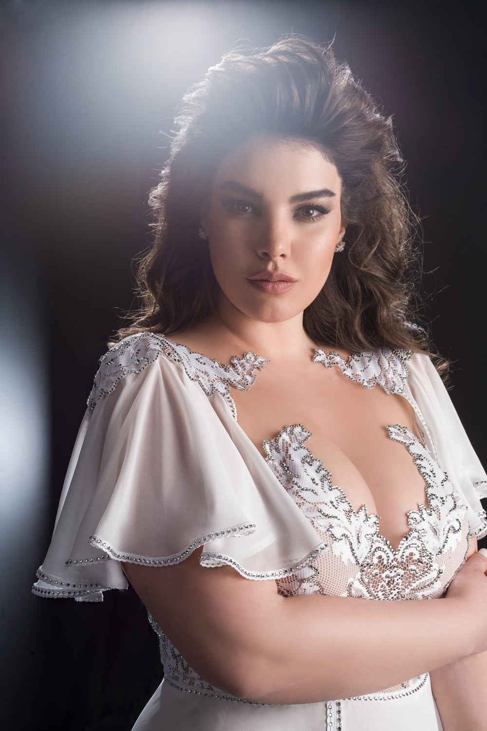 אישה עם שיער חום גלי לובשת את שמלת Harmoni מקולקציית Bridal Plus 2019, עם שרוולים שקופים ומתנפנפים וקישוטי חרוזים מורכבים. היא עומדת בביטחון עם זרועות שלובות על רקע מואר בעדינות.