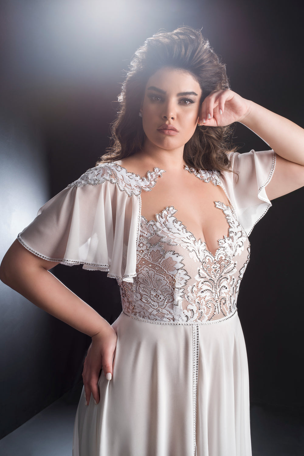 אישה בשמלת Harmoni מקולקציית Bridal Plus 2019 של דרור קונטנטו, עם תחרה לבנה, שיפון, רקמה ושרוולים שקופים מתנפנפים, פוזת בביטחון על רקע כהה, כשידה האחת מונחת על מותנה.