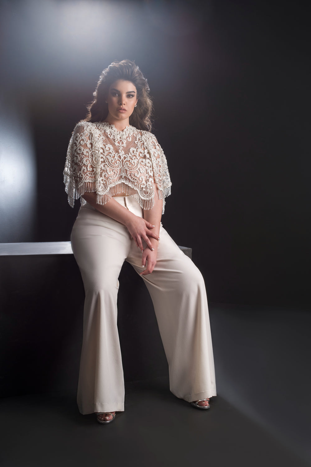 אישה עם שיער ארוך וגלי יושבת על ספסל על רקע כהה, לבושה בביטחון במכנסיים רחבים בצבע שמנת ובחולצת גלימה עם שרוולים קצרים ופרחים מתחרת Bridal Plus Collection 2019 - Alianna.