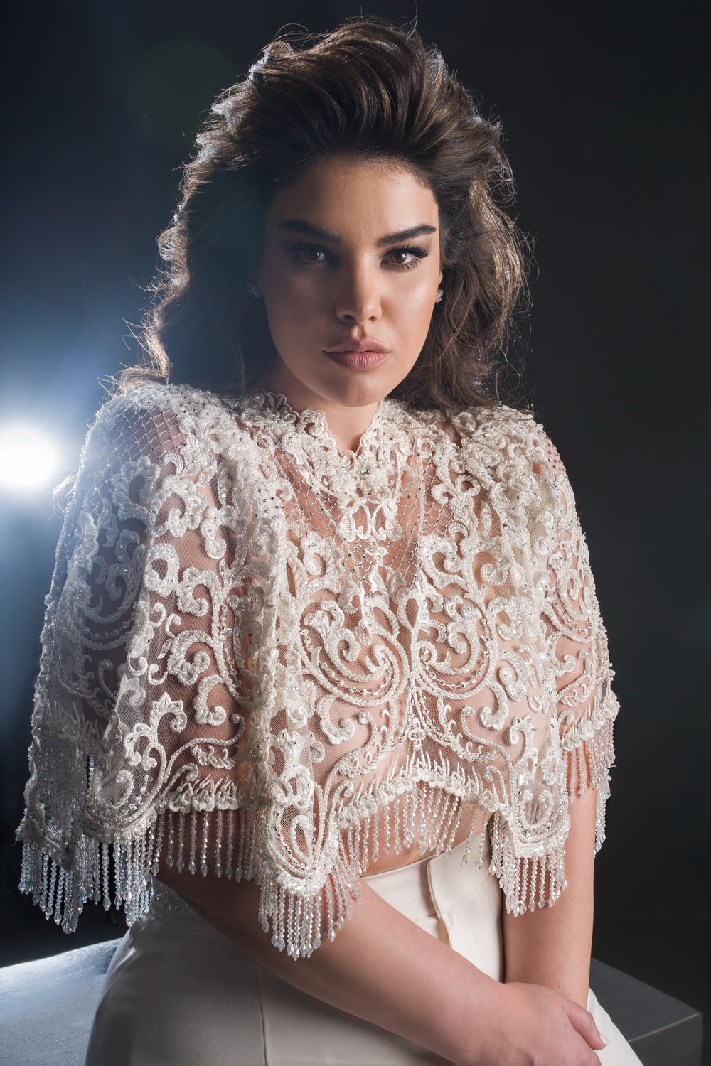 אישה עם שיער כהה ושופע מדגמנת את גלימת "אליאנה" המעוטרת והשקופה מקולקציית הכלות פלוס 2019 של דרור קונטנטו, המעוטרת בחרוזים ובפונפונים, ומצטלמת על רקע כהה המואר באור בהיר מאחוריה.