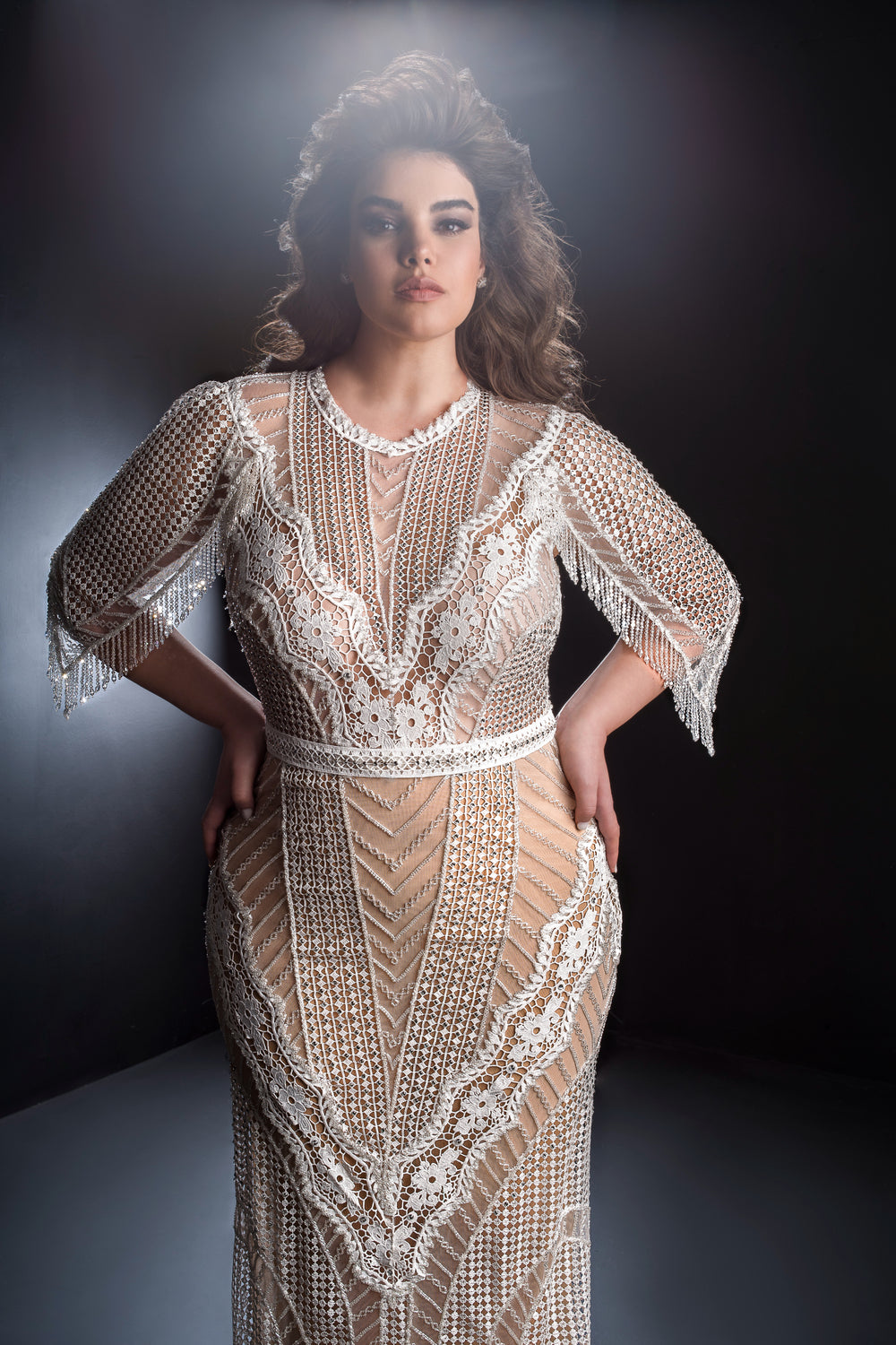 אישה עם שיער שופע מתנוססת בביטחון בשמלת Anika השקופה והרקומה מקולקציית Dror Kontento Bridal Plus 2019, עם דוגמאות גיאומטריות ותחרה על גבי בטנה בצבע גוף, על רקע כהה ומואר באור רך.